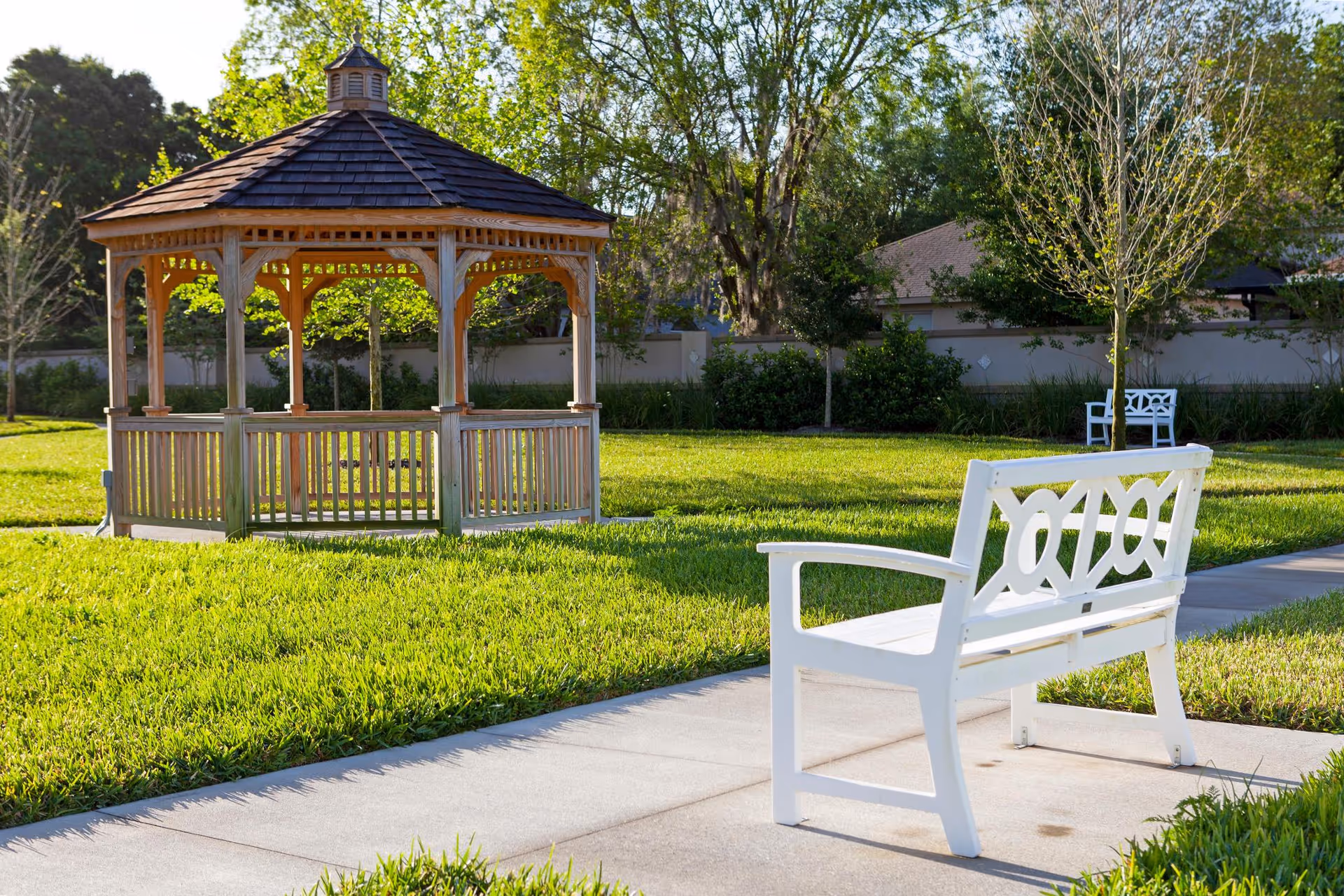 White bench on a paved path facing a wooden gazebo in a sunny grassy outdoor garden.