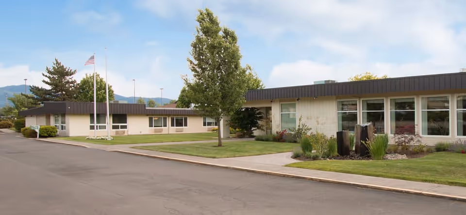 Exterior view of a single-story senior living facility building with large windows, a well-maintained lawn, trees, and an American flag on a flagpole under a partly cloudy sky.
