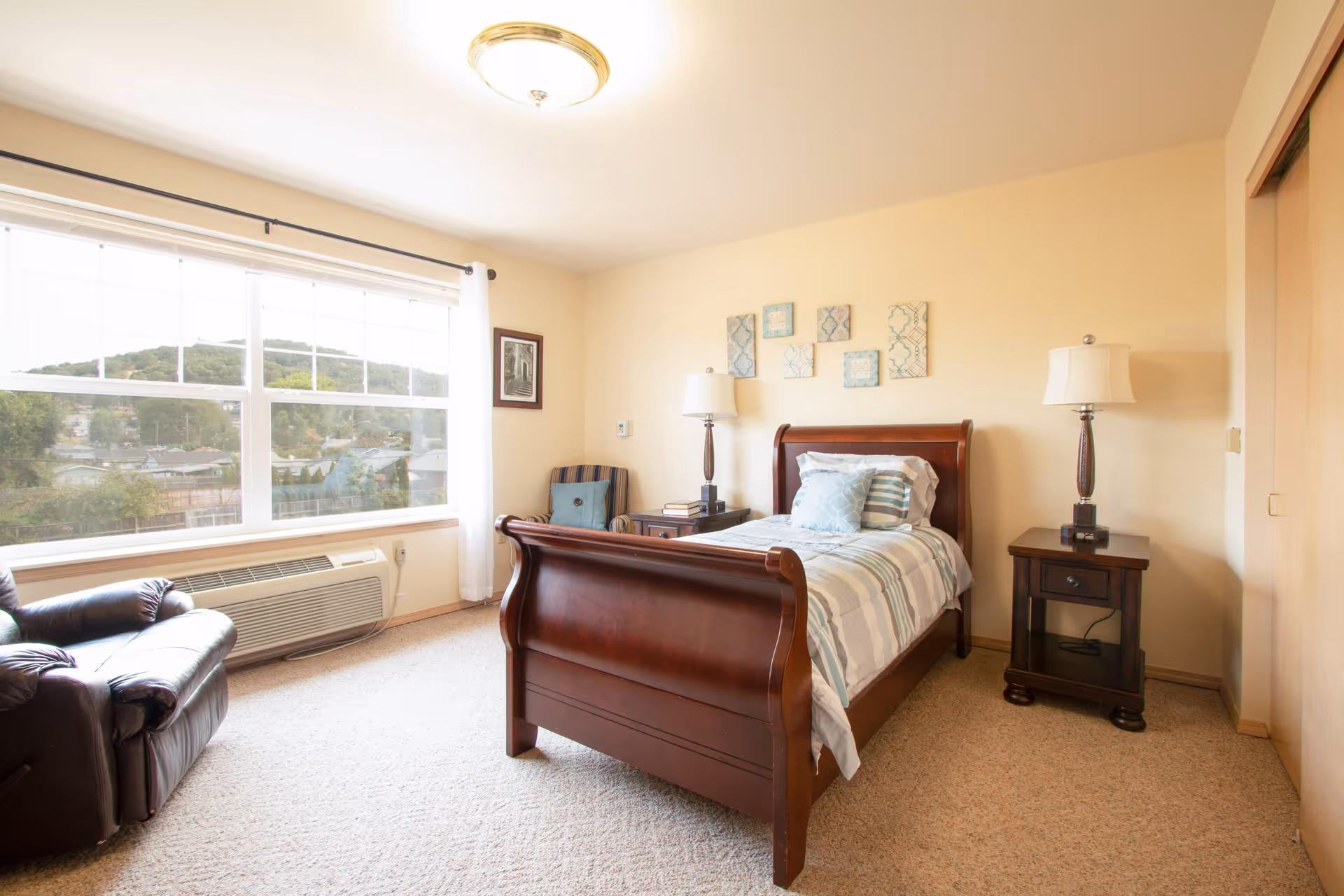 A bright and cozy bedroom with a large window showing a scenic view of trees and hills. The room features a wooden sleigh bed with striped bedding and multiple pillows, two matching wooden nightstands with lamps, a striped armchair with a cushion, a leather recliner chair, and beige carpeted flooring. The walls are painted light beige and decorated with framed artwork above the bed.