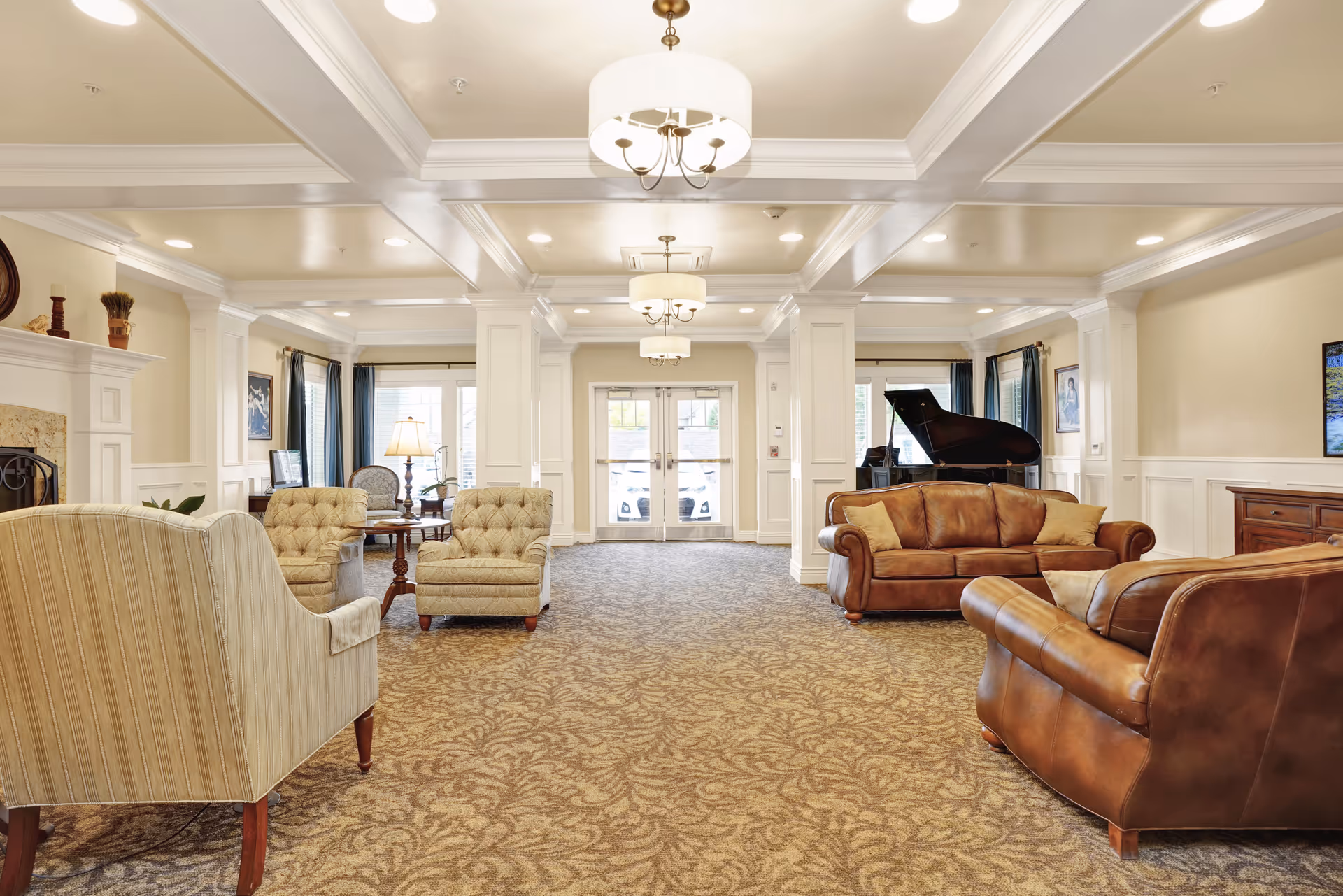 A spacious and well-lit living room area in a senior living facility with beige walls, patterned carpet, and white coffered ceiling. The room features multiple seating options including a striped armchair, two beige tufted chairs, and two brown leather sofas. A grand piano is positioned near the windows with blue curtains. There are decorative elements such as a fireplace, framed pictures on the walls, and a wooden sideboard. The room has large windows and glass doors letting in natural light.