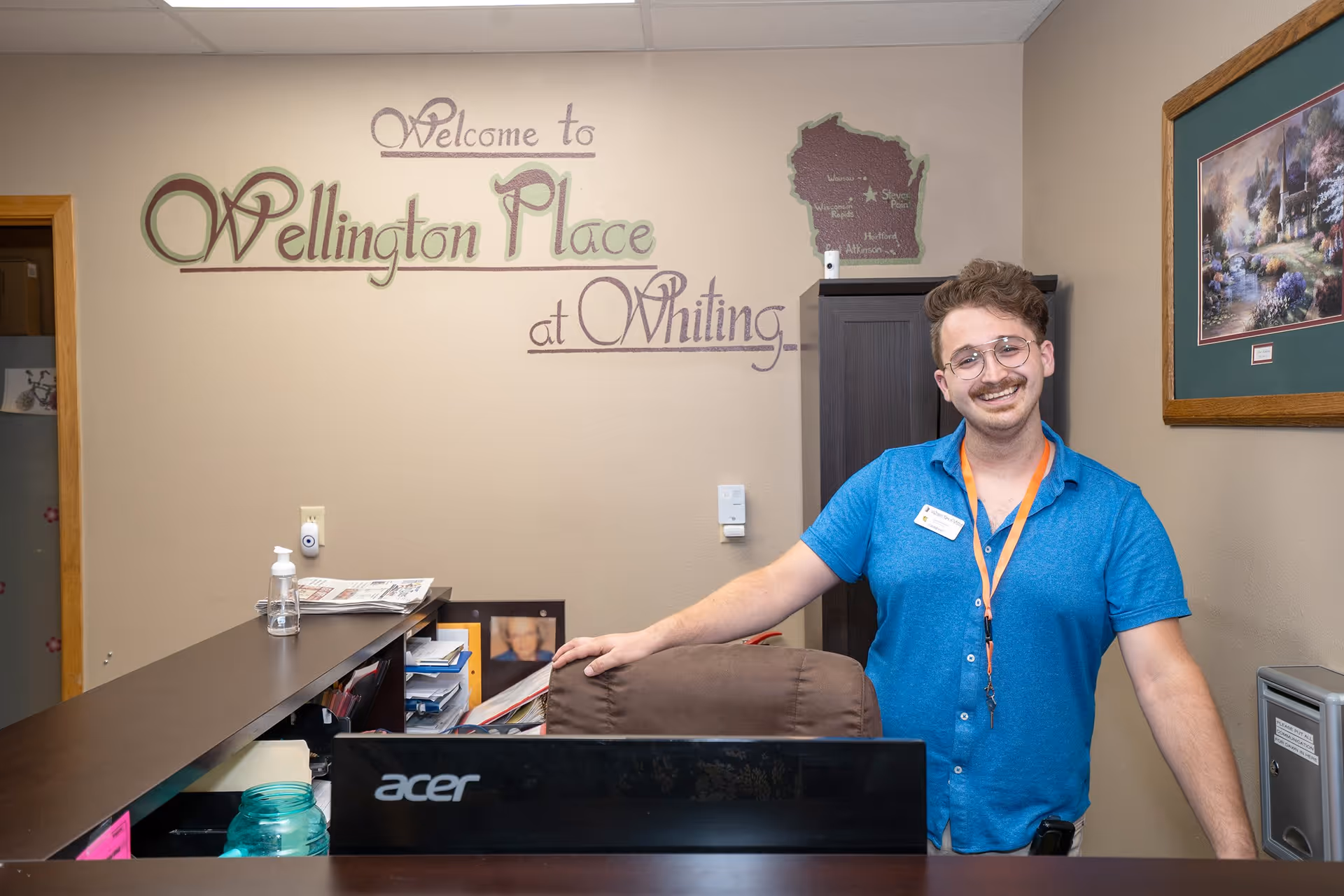 A smiling man with glasses and a mustache stands behind a reception desk in an office area. On the wall behind him is a decorative sign that reads 'Welcome to Wellington Place at Whiting.' The desk has an Acer computer monitor, a hand sanitizer bottle, and some papers. There is a framed picture on the wall to the right and a small cabinet behind the man.