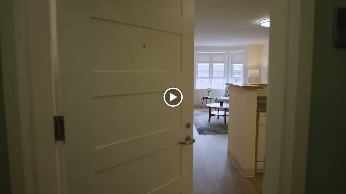 Open entry door looking into a bright living area with an armchair, coffee table, rug and a kitchen counter near bay windows.