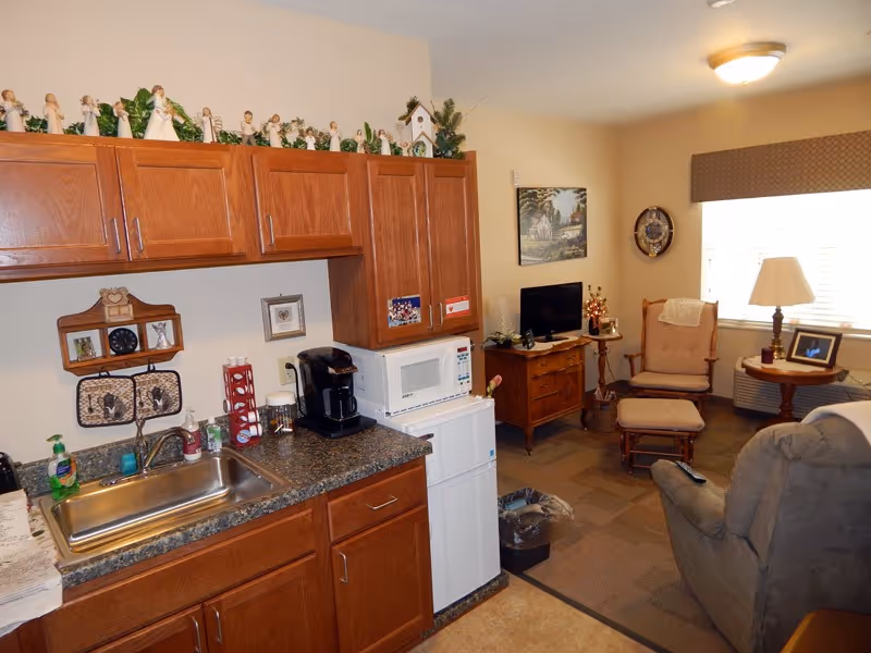 A small senior living studio interior with a kitchenette and sink in the foreground and a sitting area with recliner, TV and armchair by a window.