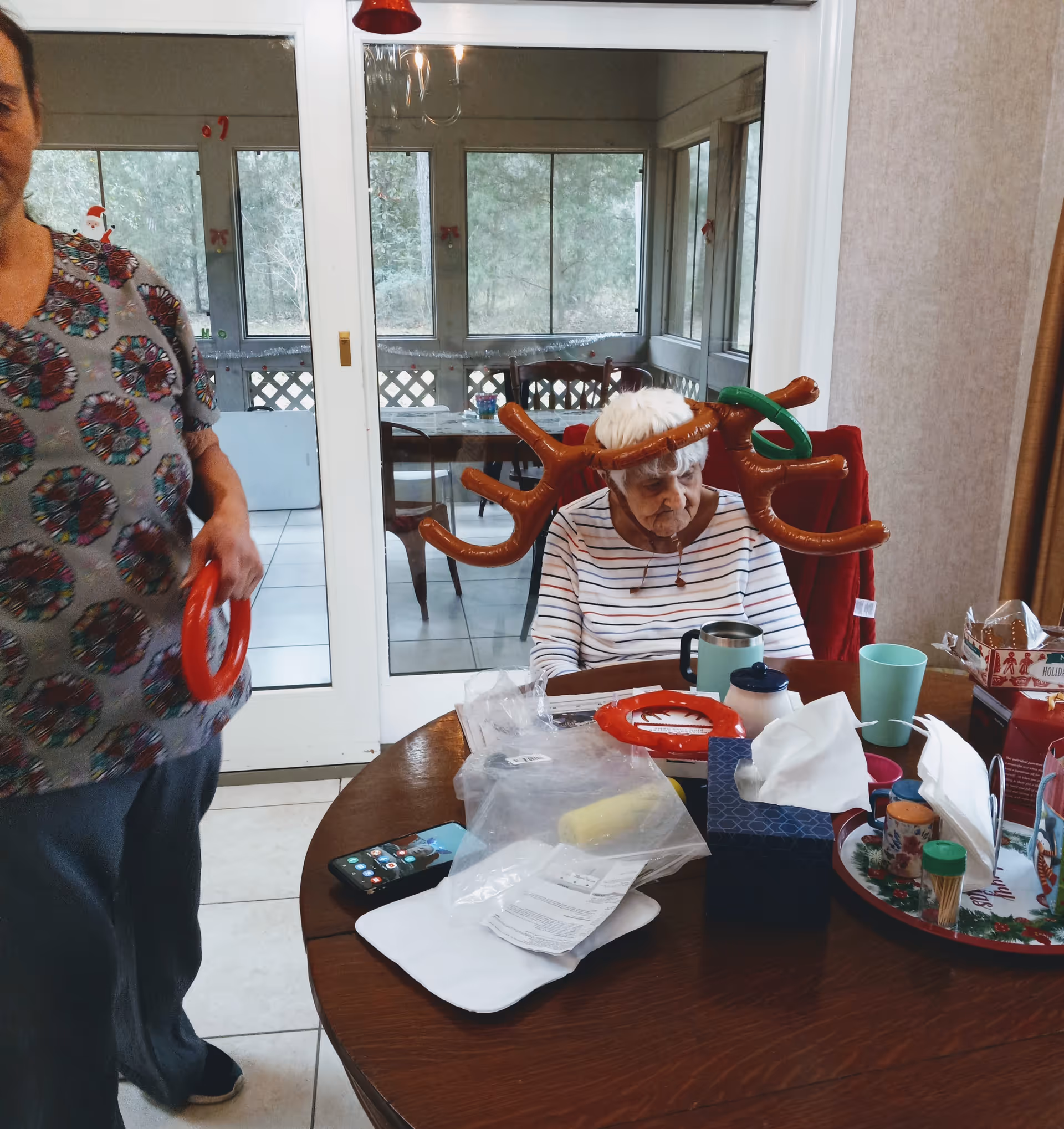 An elderly woman wearing inflatable reindeer antlers sits at a cluttered dining table while another person stands nearby holding a red inflatable ring in a dining/sunroom area.