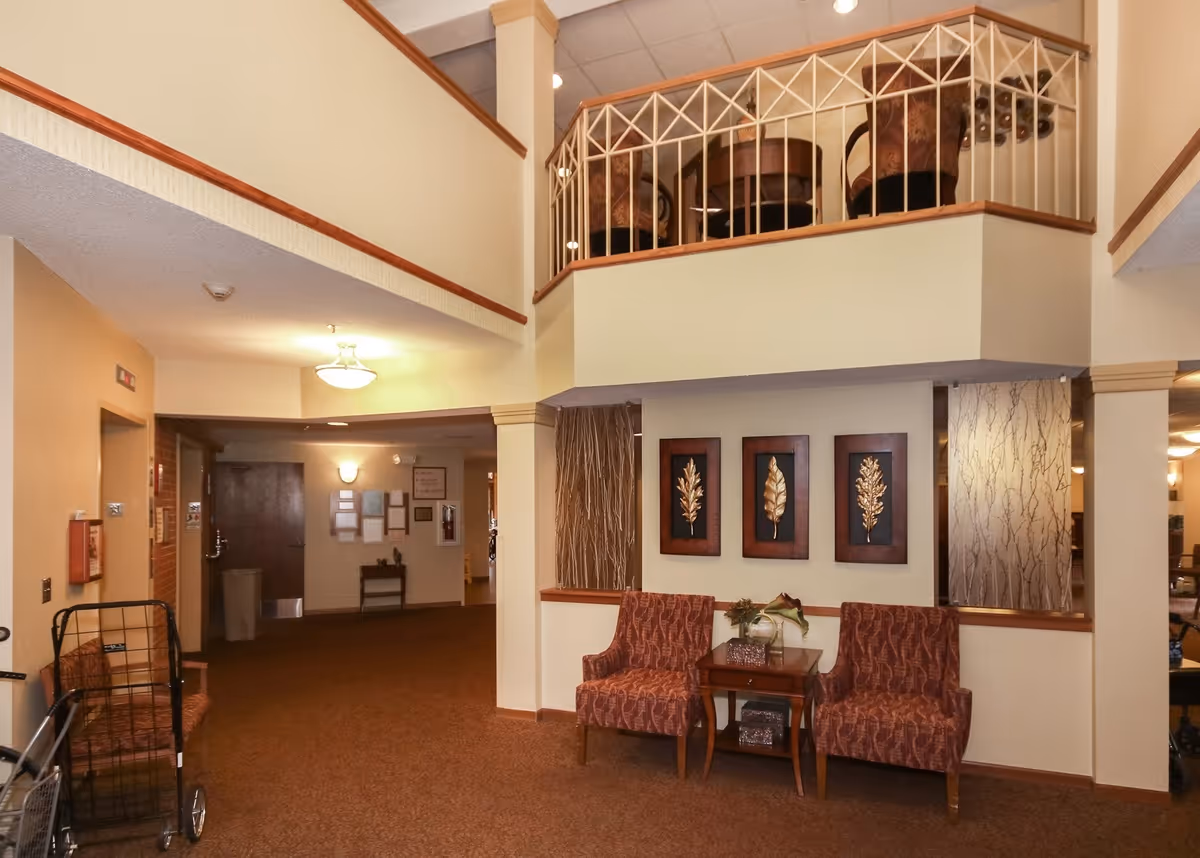 Lobby-style interior with two patterned armchairs and a small table beneath framed leaf artwork and an upper balcony.