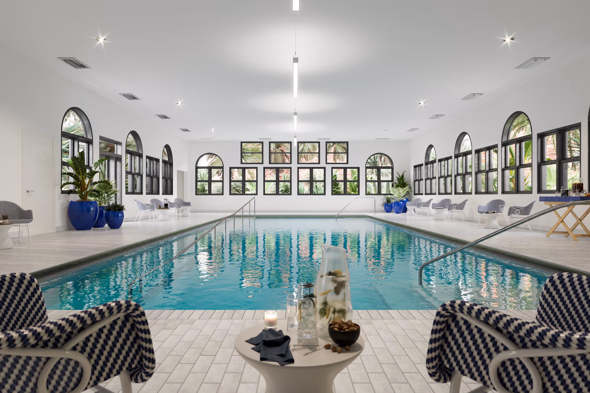 Bright indoor swimming pool room with arched windows, lounge chairs, and potted plants.