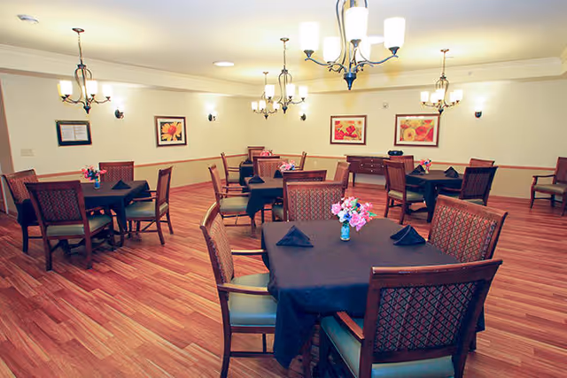 A dining room with several square tables covered with black tablecloths, each set with black folded napkins and small flower arrangements in blue vases. The room has wooden flooring, cream-colored walls with framed floral artwork, and multiple chandeliers providing warm lighting.