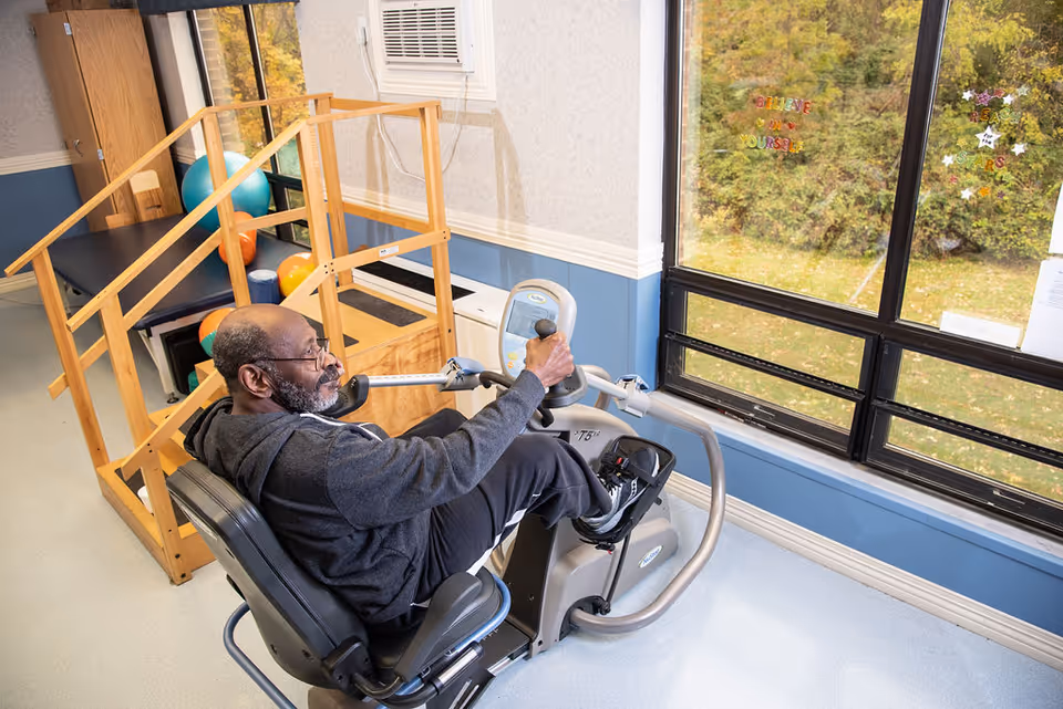 An elderly man with glasses and a beard is exercising on a seated recumbent bike in a room with large windows showing greenery outside. Behind him, there is a wooden ramp with colorful exercise balls and a padded bench. The walls are painted blue and white.