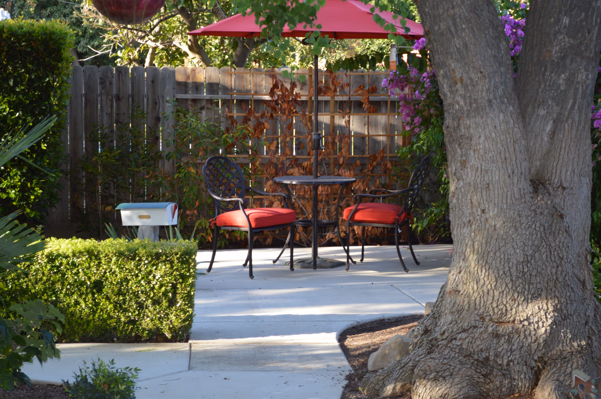 Outdoor patio area with a round metal table and two black metal chairs with red cushions under a red umbrella. The patio is surrounded by green bushes, a wooden fence, and a large tree trunk is visible in the foreground on the right side.