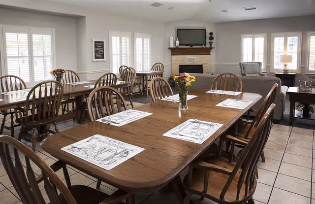 Bright communal dining room with multiple wooden tables and chairs set with placemats and flower vases, and a seating area with a fireplace and TV in the background.