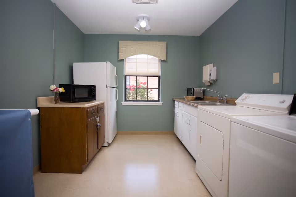 A small laundry room with green walls and beige flooring. On the left side, there is a wooden cabinet with a microwave and a small vase with flowers on top, next to a white refrigerator. On the right side, there is a white washing machine and dryer, a countertop with a sink, a toaster, and a paper towel dispenser mounted on the wall. A window with a beige valance shows some pink flowers outside.