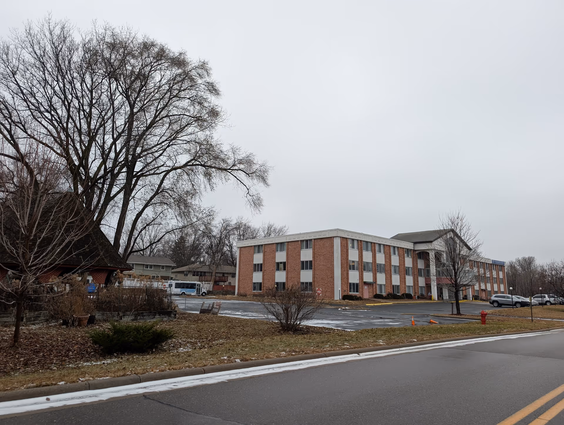 Exterior view of a three-story brick building with white accents, identified as Centennial Gardens for Nursing and Rehabilitation, on a cloudy day. There is a parking lot with several cars and a bus, leafless trees, and a small gazebo structure on the left side of the image.