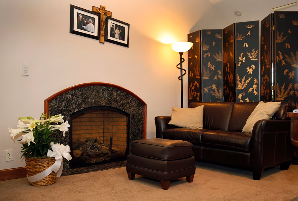 Cozy living room area with a dark brown leather sofa and matching ottoman, a floor lamp providing warm light, a decorative folding screen with gold patterns, a basket of white lilies with a white ribbon, and a fireplace with a dark granite surround. Above the fireplace are two framed photos and a wooden crucifix mounted on the wall.