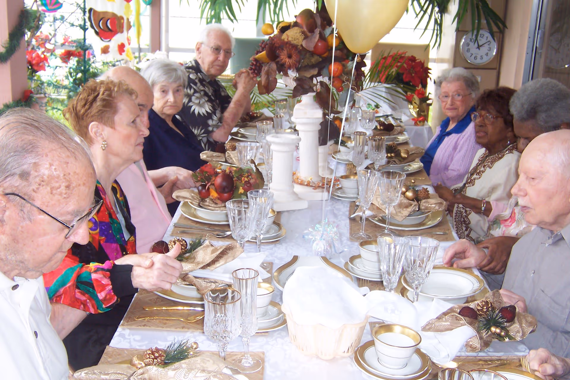 A group of elderly people seated around a long dining table set with fine china, glassware, and festive decorations including a centerpiece with autumn-themed elements and balloons. The setting appears to be a communal dining area with large windows and plants in the background.