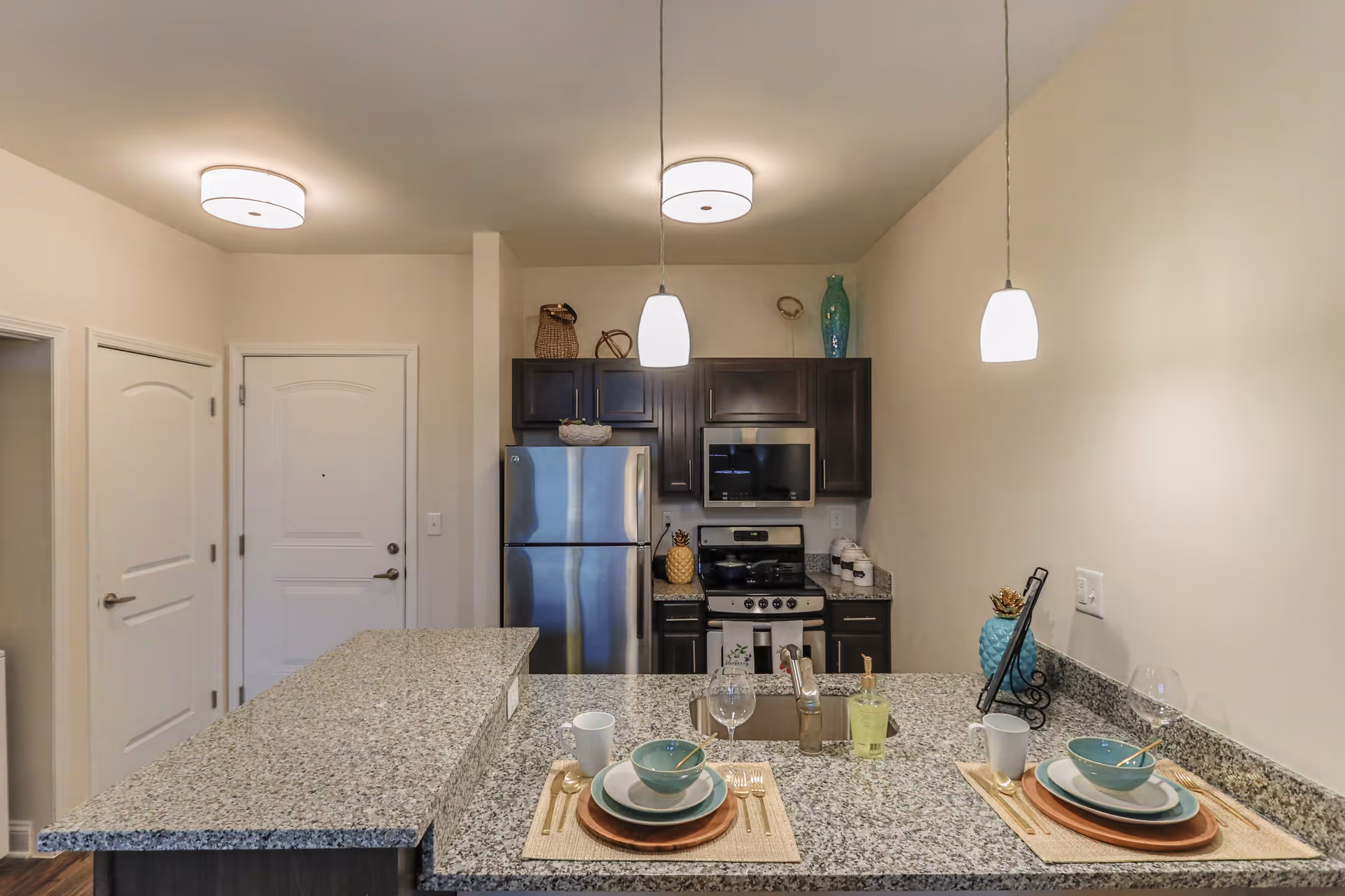 Modern kitchenette with a granite breakfast bar set for two, stainless steel refrigerator and stove, dark cabinets, and pendant lights.