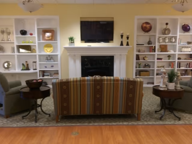 A cozy living room area with a striped sofa facing a fireplace and a wall-mounted flat screen TV above it. On either side of the fireplace are white built-in shelves filled with decorative items such as vases, plates, and small sculptures. Two armchairs flank the sofa, each next to a round wooden side table with decorative objects on top. The room has a warm, inviting atmosphere with soft yellow walls and a carpeted floor in the seating area, transitioning to wood flooring in the foreground.