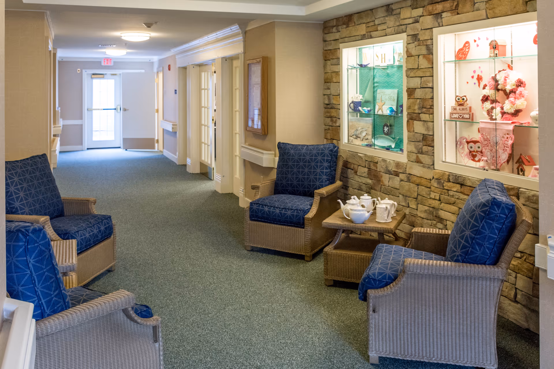 A hallway in a senior living facility with four wicker armchairs featuring blue cushions arranged around a small wicker table with a white tea set. The right wall is decorated with stone veneer and two glass display cases containing various decorative items including flowers, owls, and seashells. The hallway has a green carpet and beige walls with white trim, and a door with an exit sign is visible at the far end.