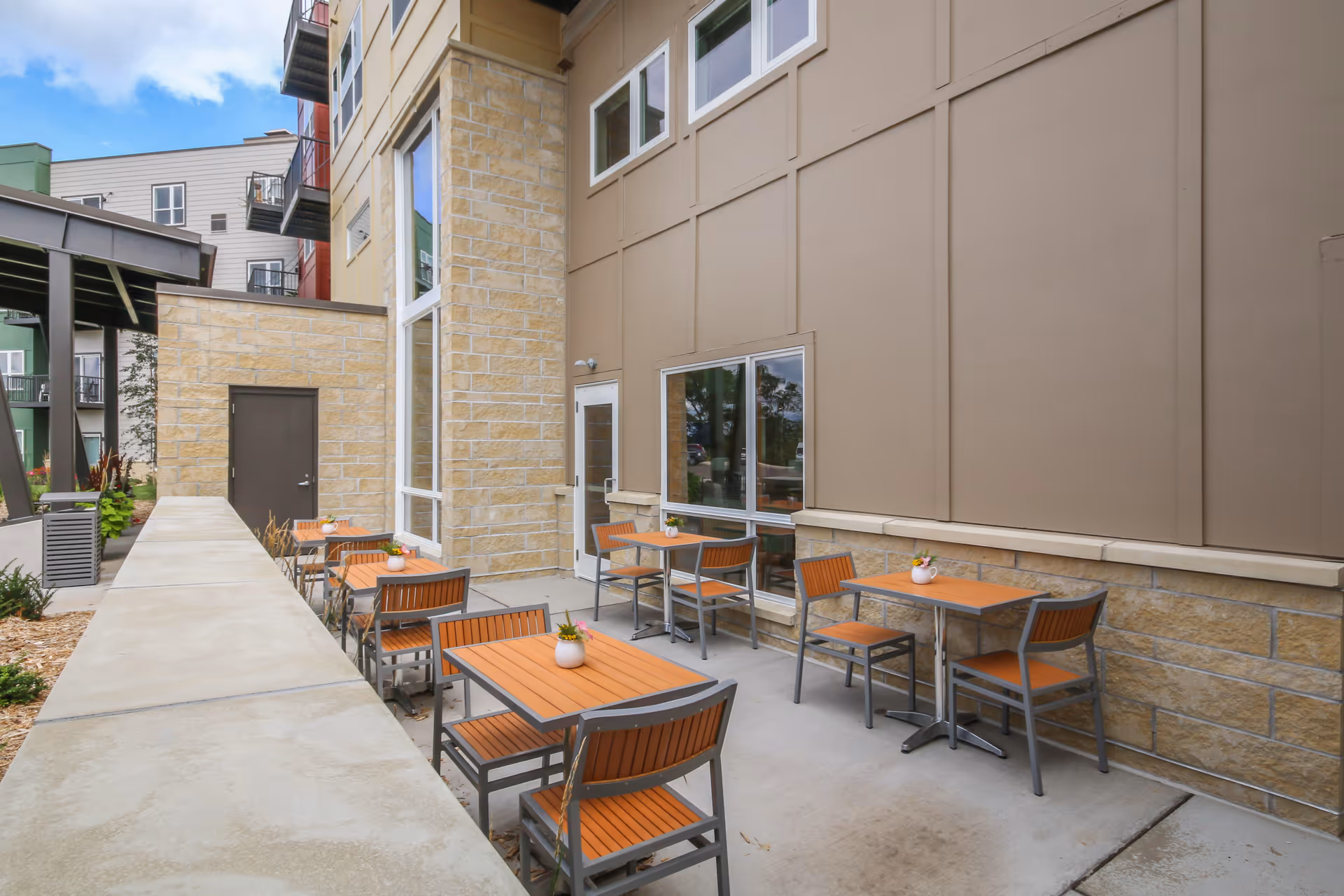 Outdoor patio with multiple small square tables and orange chairs along the exterior wall of a modern building.