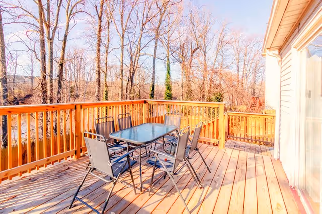 Outdoor wooden deck with a glass-top table and six metal chairs, surrounded by a wooden railing. Leafless trees are visible in the background under a clear sky.
