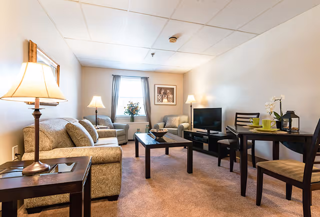 A cozy living room with beige walls and carpet, featuring a patterned sofa and armchairs, a wooden coffee table, a TV on a stand, a dining table with chairs, and lamps providing warm lighting. A window with curtains and a framed picture are visible on the far wall.