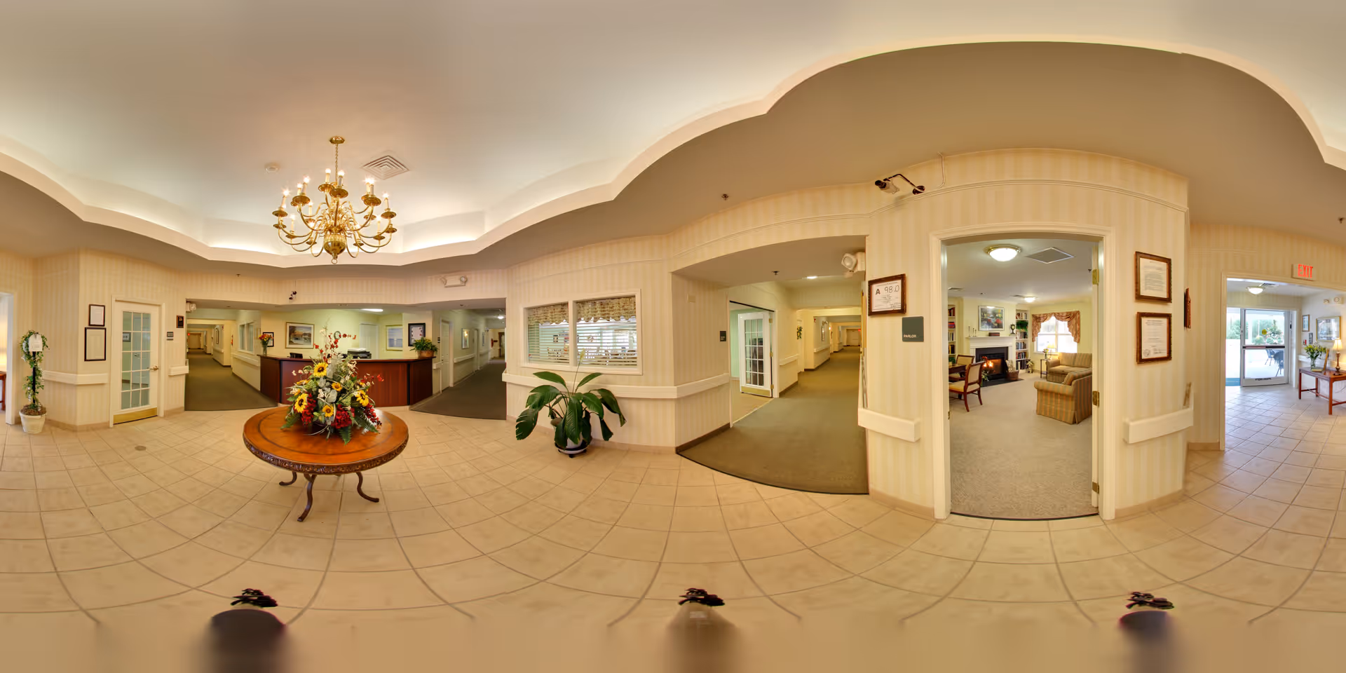 A spacious and well-lit senior living facility lobby with a round wooden table in the center holding a large floral arrangement. The area features tiled flooring, beige walls with subtle stripes, and a decorative ceiling with a chandelier. Multiple doorways and hallways lead to other parts of the facility, including a cozy sitting area with armchairs and a fireplace, and an exit door with glass panels allowing natural light to enter. Potted plants and framed pictures decorate the space.