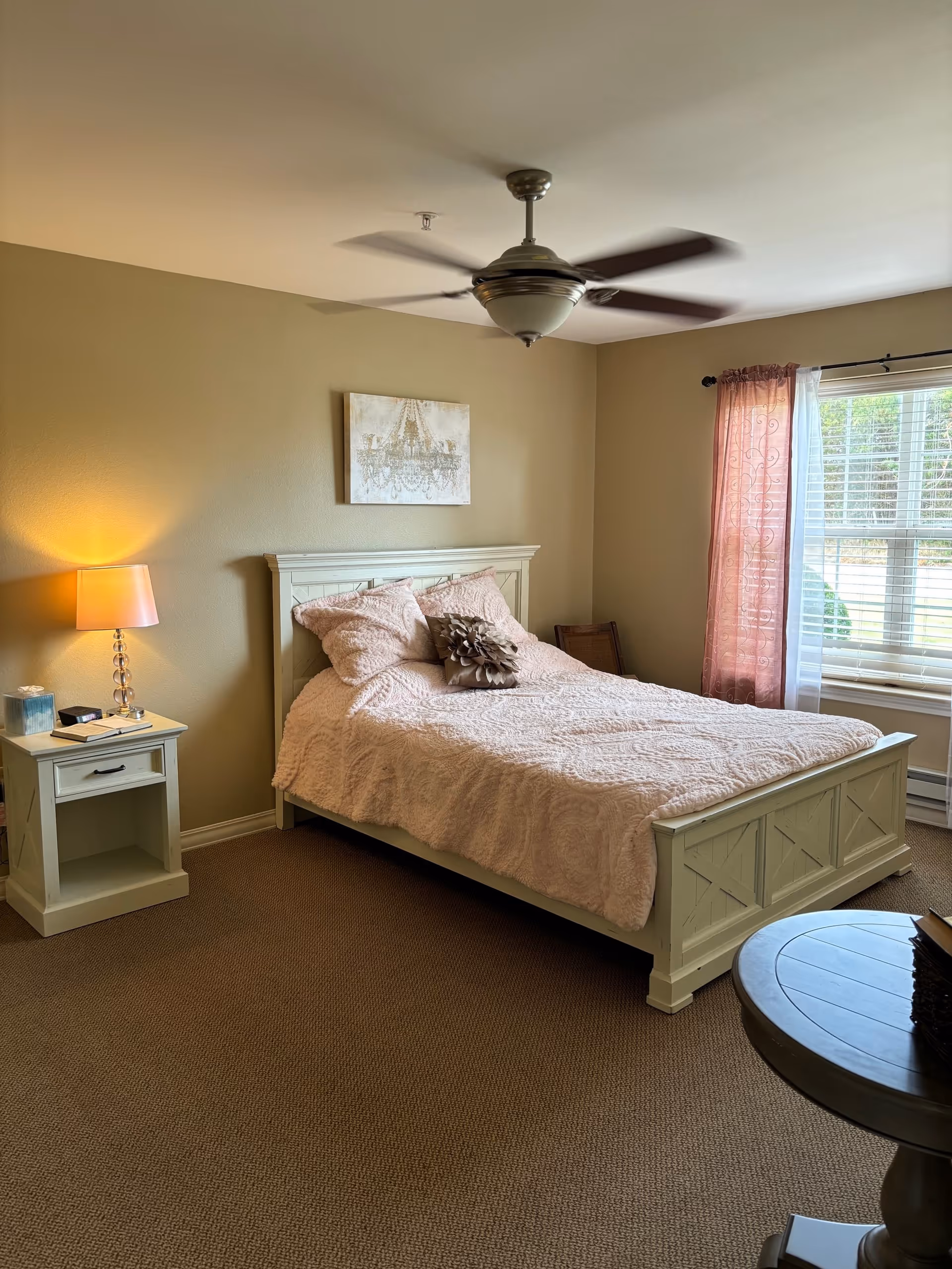 A cozy bedroom with a white wooden bed frame, a pink quilted bedspread, and several pillows. There is a nightstand with a lamp and some items on it to the left of the bed. A ceiling fan is spinning above, and a window with pink and white curtains lets in natural light on the right side of the room. A round table with books is partially visible in the foreground.