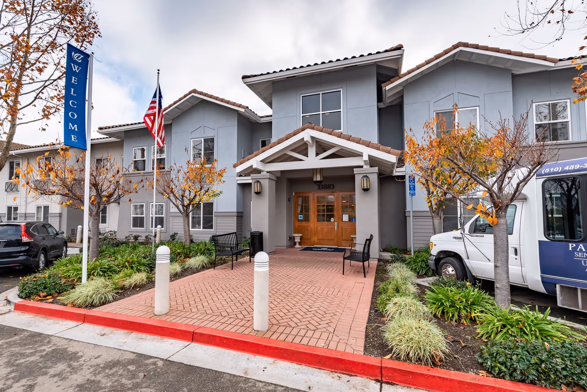 Front exterior view of a senior living facility with a covered entrance, two benches, a flagpole with an American flag, and a blue welcome banner. There are small trees with autumn leaves and parked vehicles including a white shuttle van.