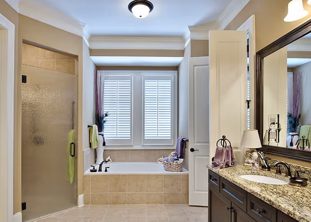 A bright and clean bathroom featuring a tiled bathtub beneath two shuttered windows, a glass-enclosed shower with a towel hanging on the door, and a granite countertop with a sink, mirror, and various toiletries.