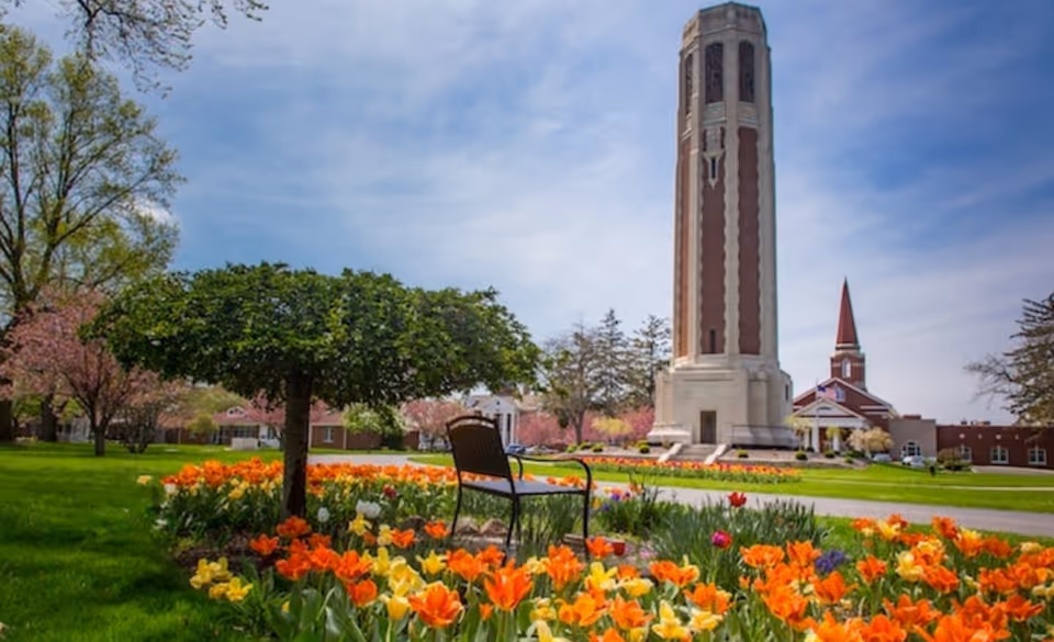 A landscaped lawn with orange and yellow tulips, a chair under a small tree, and a tall brick tower and church steeple in the background under a blue sky.