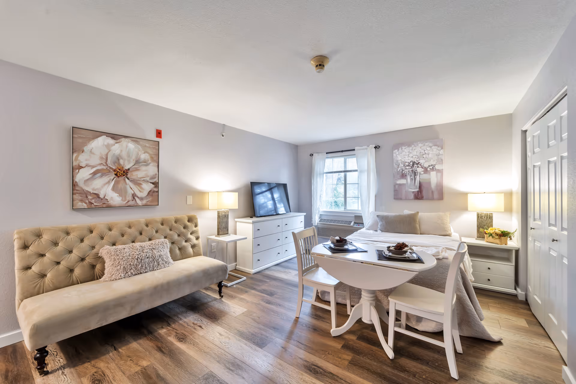 A bright and cozy senior living room featuring a beige tufted sofa with a decorative pillow, a white dresser with a flat-screen TV on top, a small round dining table set for two, and a bed with neutral bedding. The room has wooden flooring, two table lamps on white nightstands, floral wall art, and a window with white curtains letting in natural light.