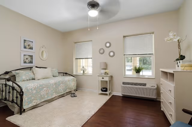 Sunlit, simply furnished bedroom with a daybed, nightstand, dresser, two windows, and a ceiling fan.