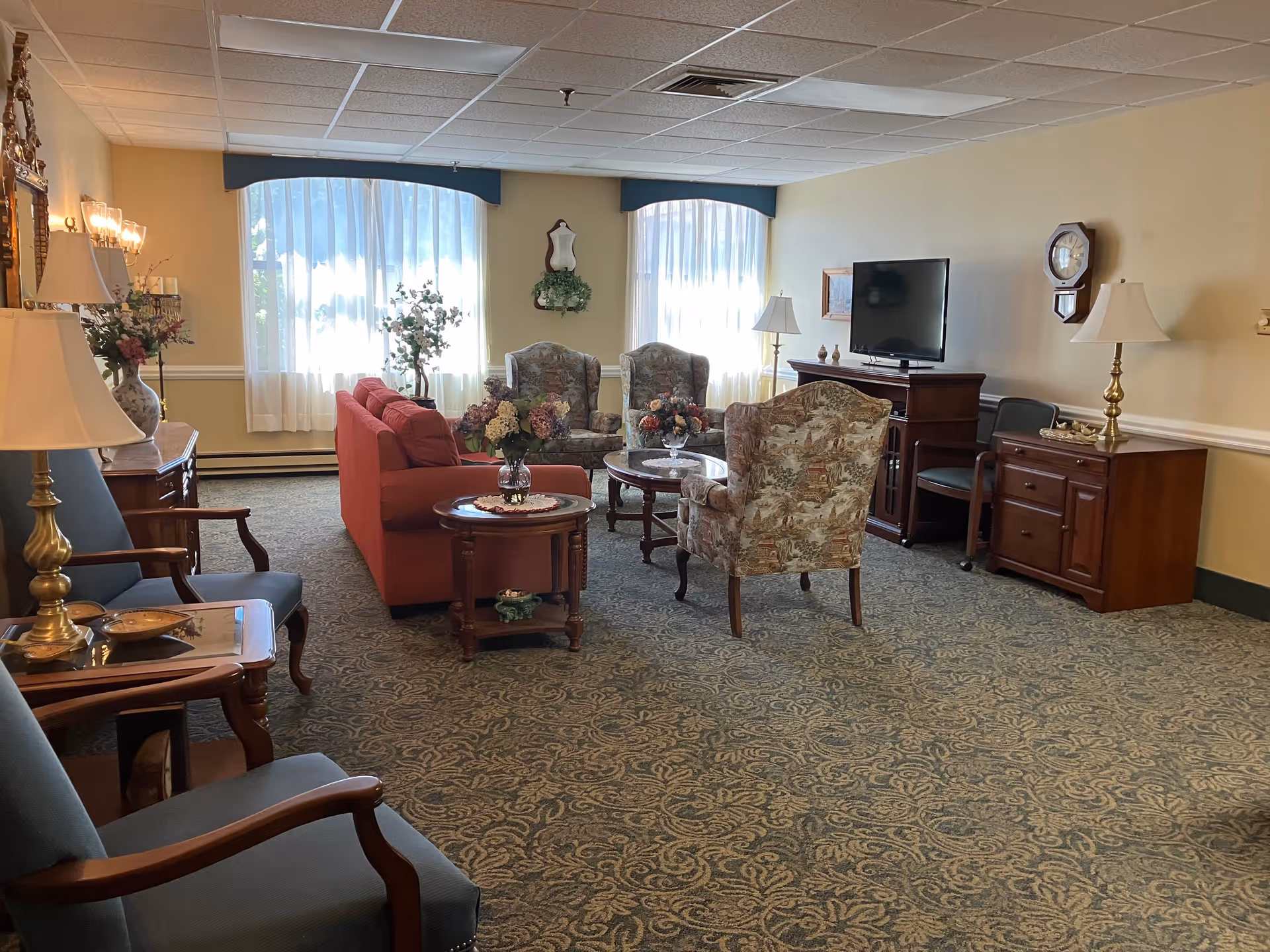 A cozy living room area with patterned carpet, two large windows with sheer white curtains, a red sofa, two floral armchairs, a glass coffee table with flower arrangements, a TV on a wooden cabinet, and additional wooden furniture including side tables and lamps.