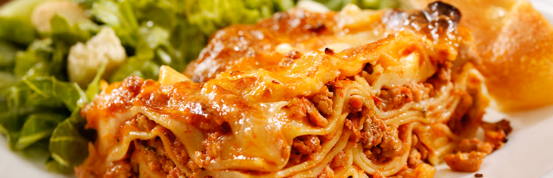 Close-up of a plate with a serving of lasagna topped with melted cheese, accompanied by a side of green salad and a bread roll.