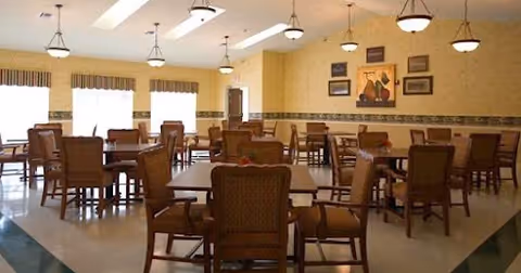 A spacious dining room with multiple wooden tables and chairs arranged neatly. The room has large windows with valance curtains allowing natural light to fill the space. The walls are painted a warm yellow with decorative trim and framed artwork. Ceiling lights hang evenly spaced across the room.