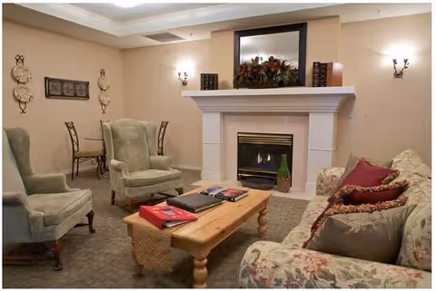 A cozy living room with a floral patterned sofa adorned with multiple pillows, two green upholstered armchairs, a wooden coffee table with books on it, and a fireplace with a mantel decorated with books and a floral arrangement. The walls are beige with wall sconces providing soft lighting, and a small dining table with chairs is visible in the background.