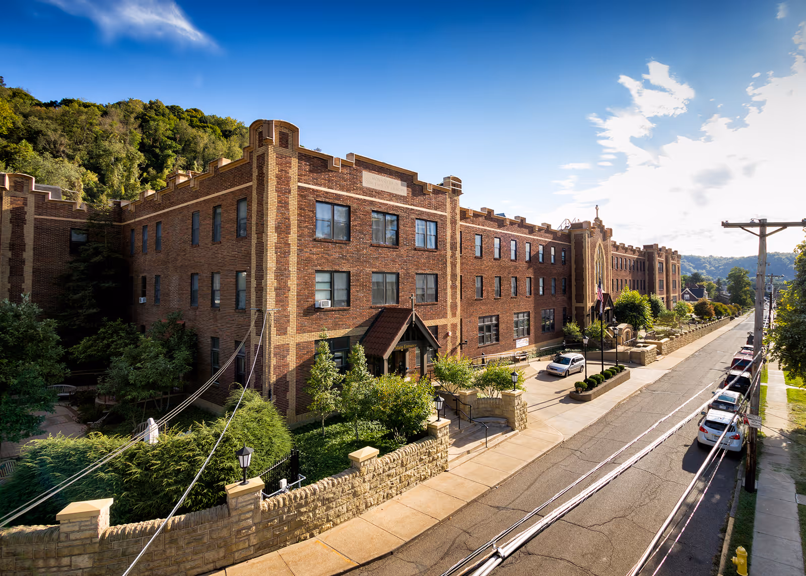 Exterior view of a large brick building with multiple windows, surrounded by greenery and a stone fence. The building is located on a street with parked cars and utility poles, under a partly cloudy blue sky.