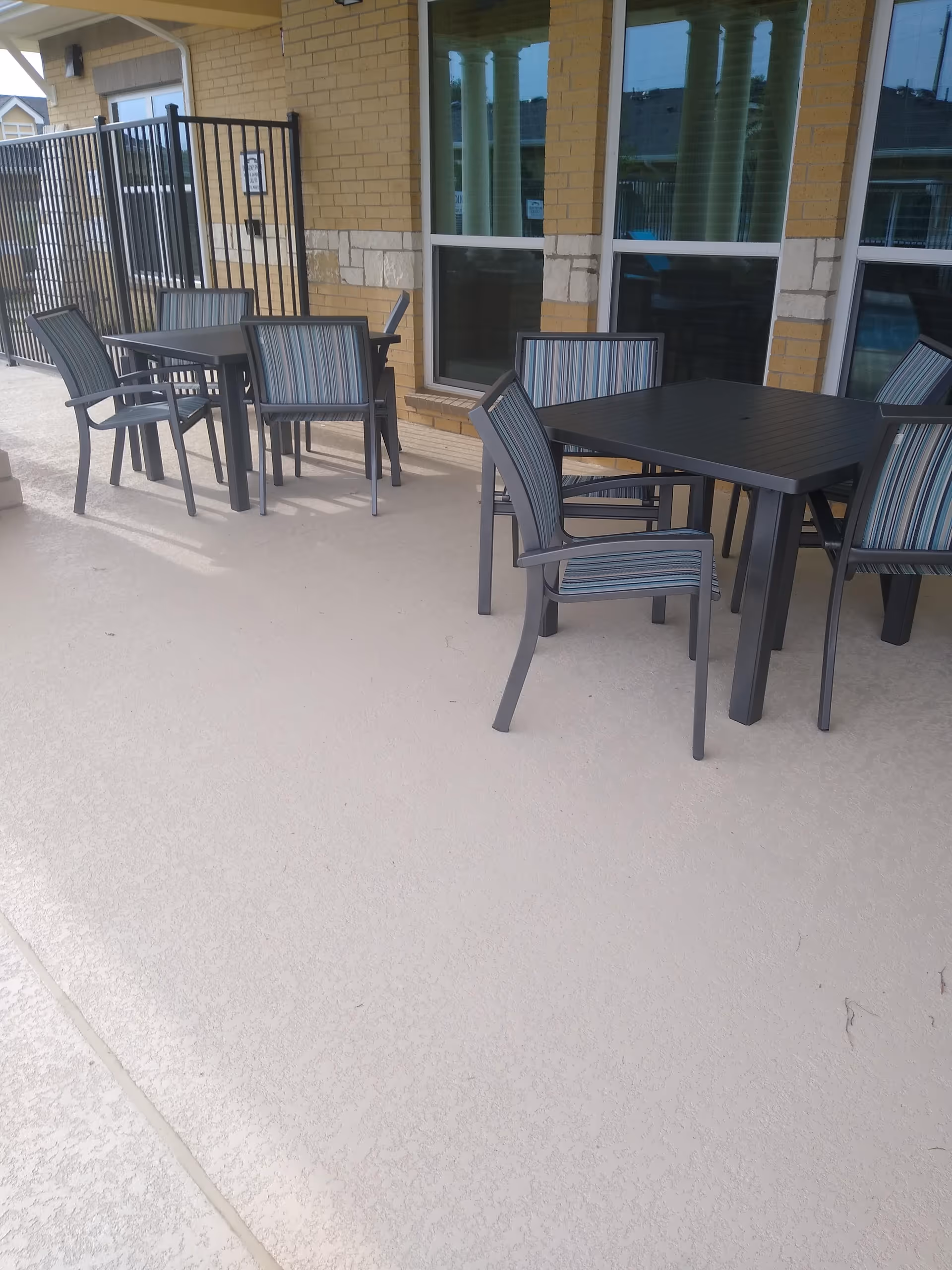 Outdoor patio area with two black square tables and several striped cushioned chairs arranged around them. The patio is adjacent to a building with yellow brick walls and large windows.