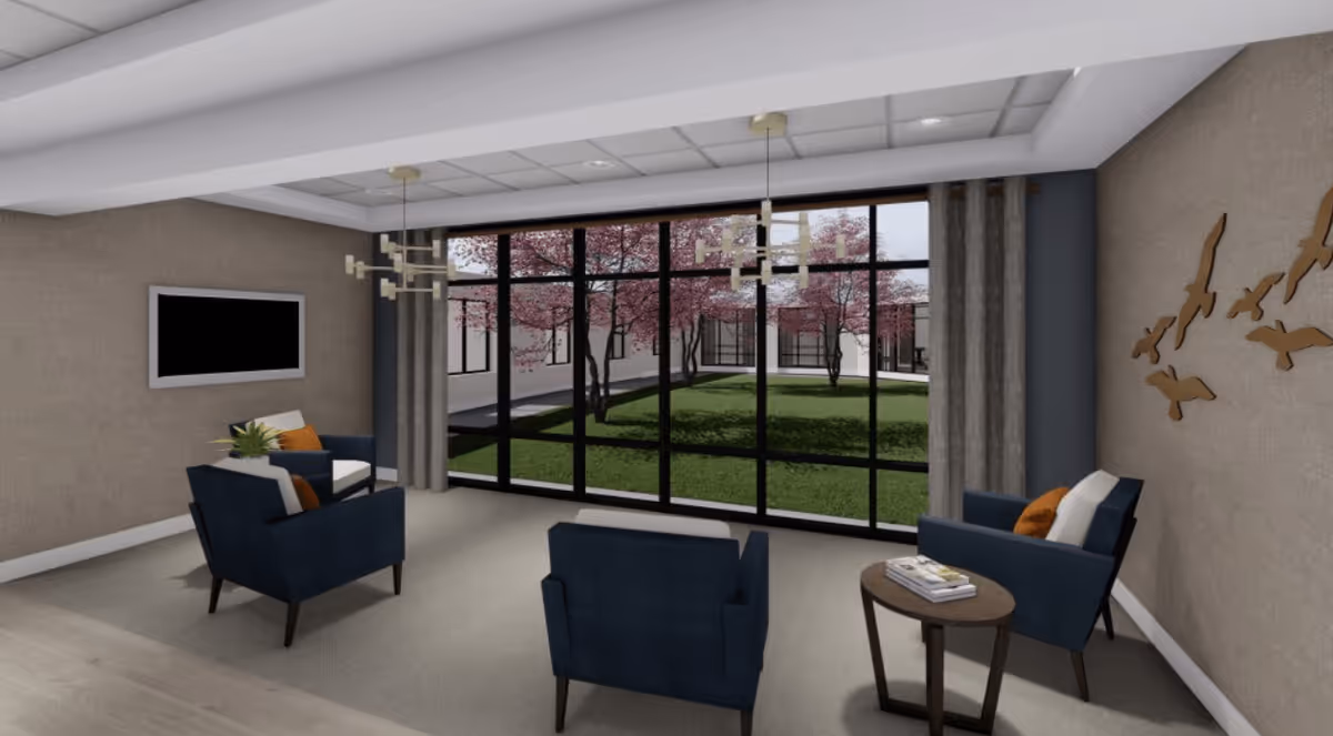 A modern lounge area with four navy blue armchairs arranged around a small round wooden table with books on it. The room has large floor-to-ceiling windows looking out onto a courtyard with green grass and pink blossoming trees. The walls are beige with a decorative wall art of birds on one side and a flat-screen TV mounted on another. Two contemporary chandeliers hang from the ceiling.