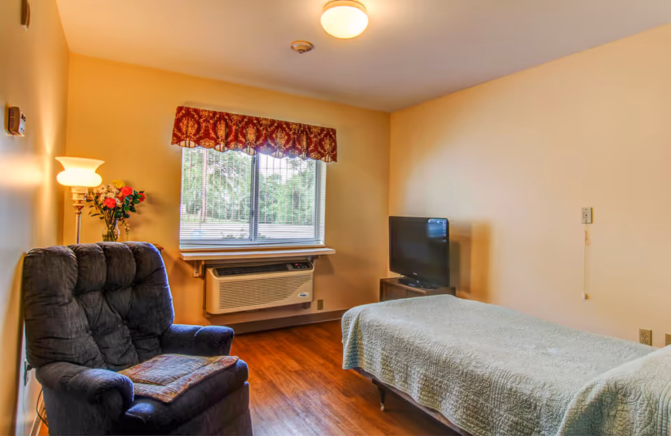 A modest senior living bedroom with a single bed covered by a green quilt, a dark upholstered recliner, a TV on a stand, a window with an air conditioner, and a floor lamp.