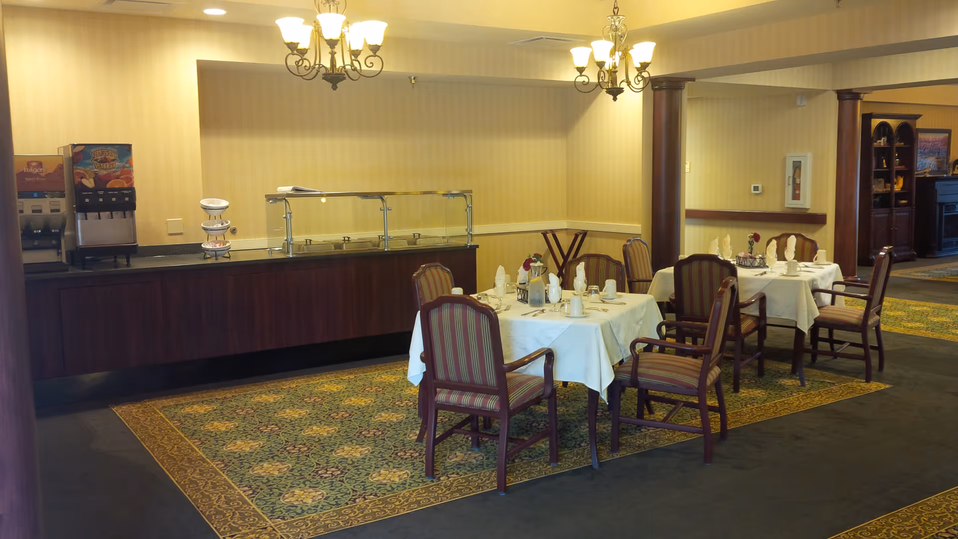Dining area with tables set for meals, upholstered chairs, a buffet counter with beverage dispenser, and chandeliers overhead.