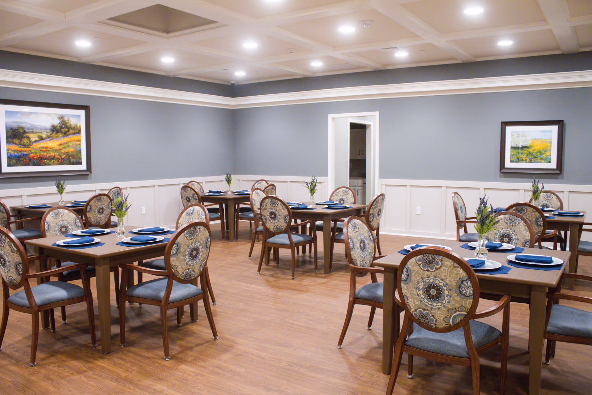 A dining room with several wooden tables and chairs arranged neatly. Each table is set with blue napkins, white plates, and small vases with flowers. The walls are painted gray with white wainscoting, and two framed landscape paintings hang on the walls. The room has a wooden floor and recessed ceiling lights.