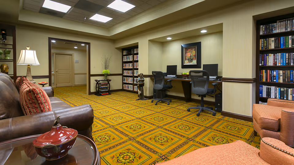 A cozy senior community room with a patterned yellow carpet, two computer workstations with chairs, bookshelves filled with books, a brown leather sofa with striped pillows, an orange armchair, a side table with a decorative red ceramic container, a lamp, and a framed artwork on the wall.