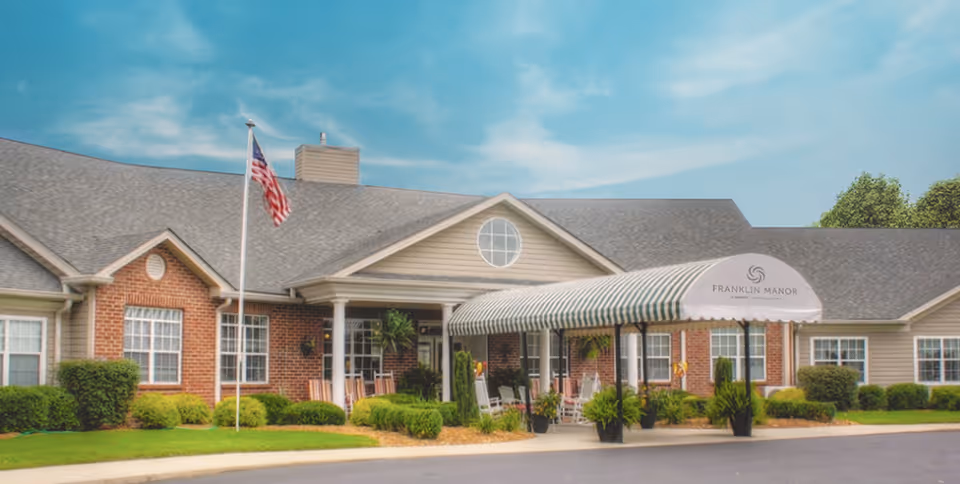 Front exterior view of Franklin Manor, a single-story building with a brick and siding facade, a covered entrance with a striped awning, several rocking chairs on the porch, an American flag on a flagpole, and well-maintained landscaping with bushes and grass.