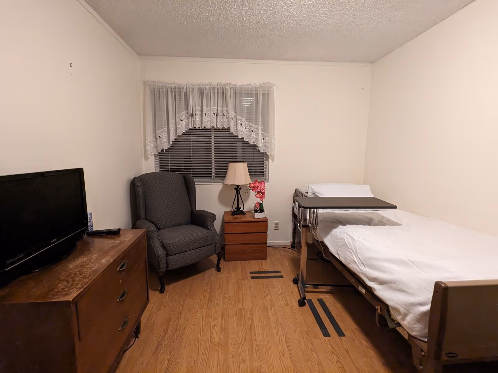 Small furnished bedroom with a single medical bed and overbed table, armchair, dresser with TV, and a nightstand under a curtained window.