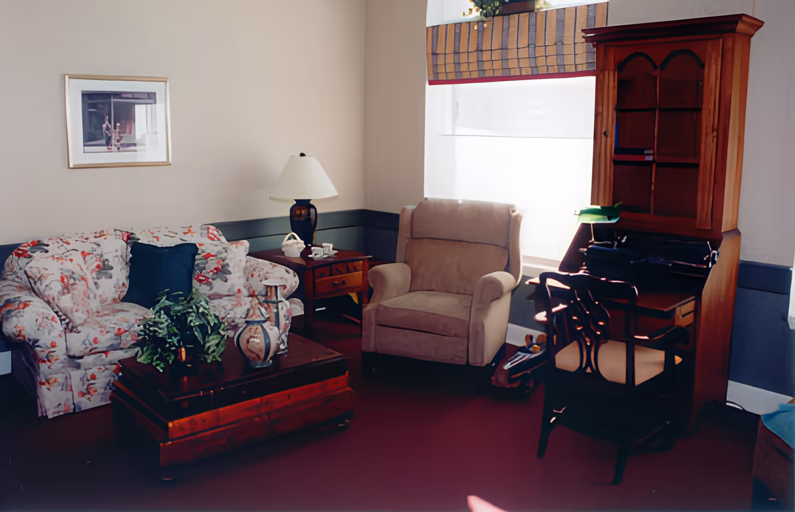 A cozy living room with a floral-patterned sofa, a beige armchair, a wooden coffee table with a plant and a vase, a side table with a lamp and a basket, a wooden desk with a chair, and a window with a striped valance.
