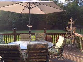 Outdoor wooden deck with a round glass table surrounded by six cushioned chairs. A large beige patio umbrella is centered over the table. In the background, there is a green lawn and dense trees.