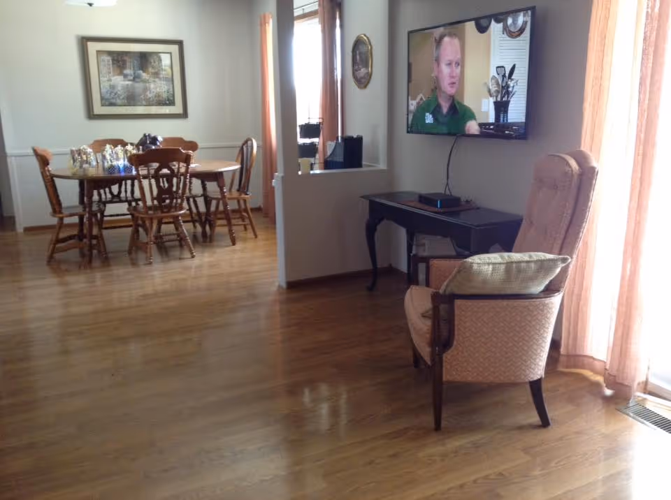 A cozy interior space with wooden flooring featuring a dining area with a wooden table and chairs, and a sitting area with an upholstered armchair and a small black table. A flat-screen TV is mounted on the wall showing a man speaking. Light peach curtains cover the windows, and a framed picture hangs on the wall above the dining table.