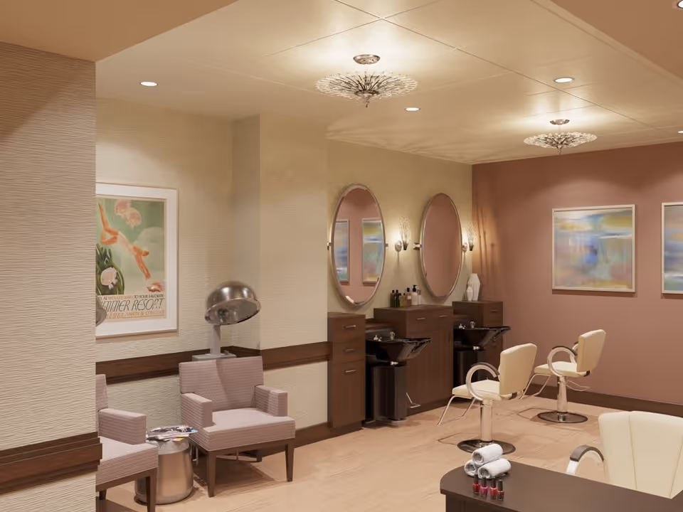 Interior view of a salon area in a senior living facility featuring two styling stations with mirrors and sinks, two salon chairs, a hair drying chair with a hood dryer, and comfortable seating. The walls are decorated with framed artwork and the lighting includes ceiling fixtures and wall sconces.