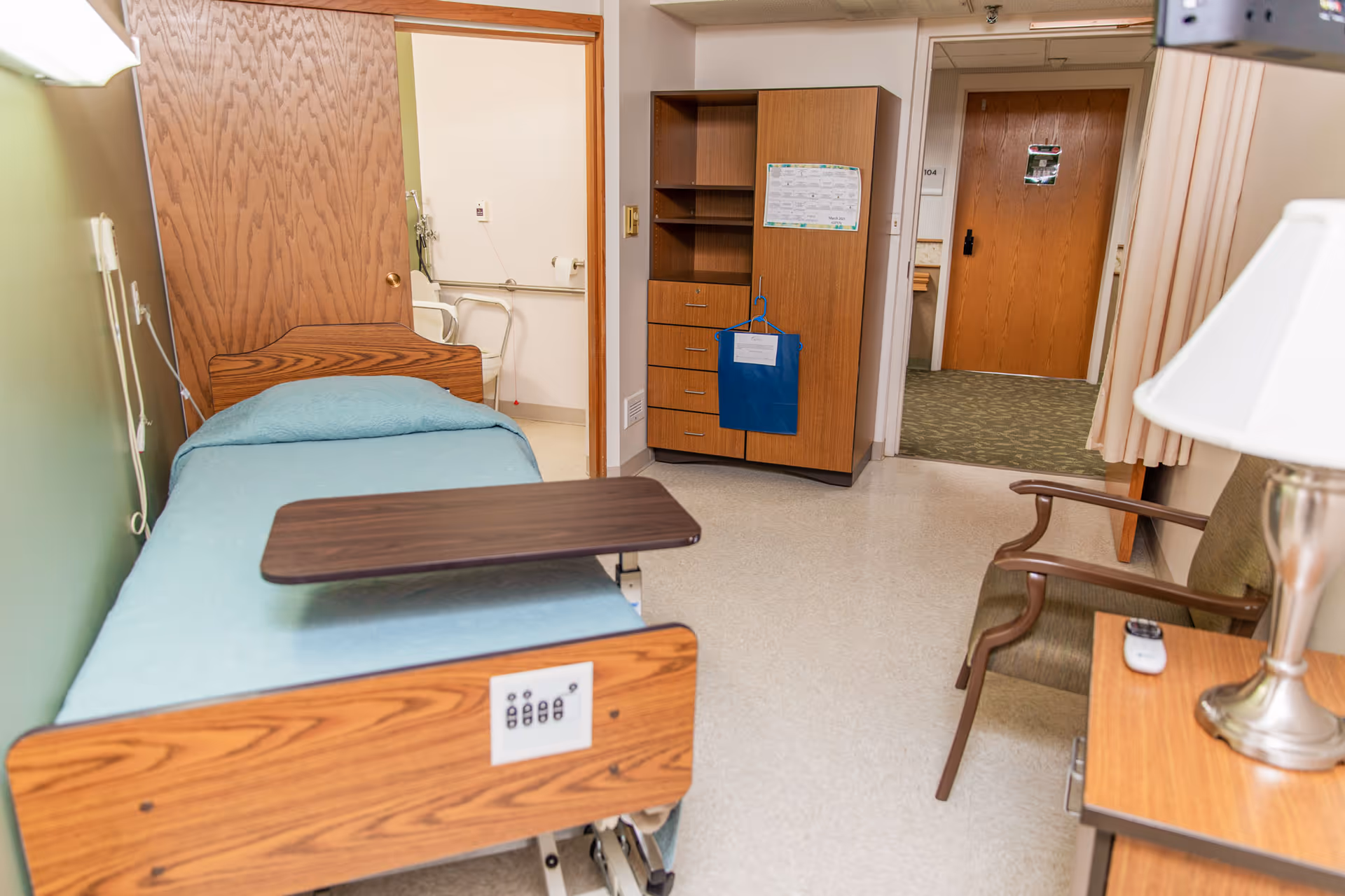 A senior living facility room with a single bed covered in a light blue blanket, a wooden adjustable overbed table, a wooden cabinet with drawers and shelves, a green armchair, and a small wooden side table with a lamp and remote control. The room has a door leading to a bathroom with grab bars and a toilet paper holder visible, and another door leading to a hallway with carpeted flooring.