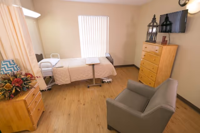 A senior living facility bedroom with a hospital-style bed covered in a beige quilt, a small overbed table, a gray armchair, a wooden dresser with decorative items including a lantern and a letter L, a wall-mounted TV, a clock, and a window with vertical blinds letting in natural light.